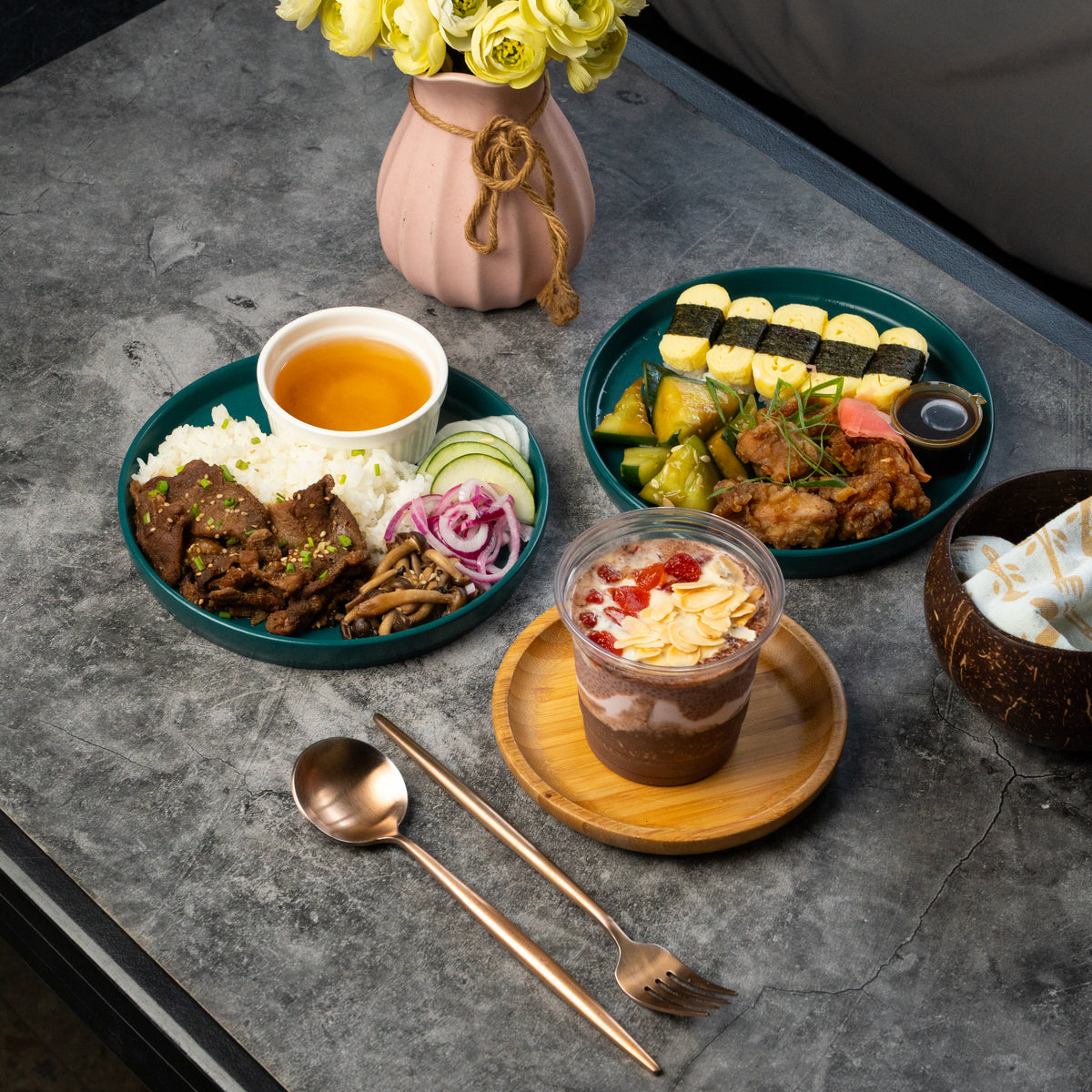 A styled food spread on a gray stone tabletop, featuring two teal plates of prepared rice and meat, a small cup of orange drink, a layered dessert in a clear cup on a wooden saucer, a pink vase with yellow flowers, and cutlery in the foreground.