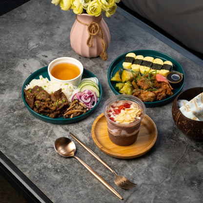 A styled food spread on a gray stone tabletop, featuring two teal plates of prepared rice and meat, a small cup of orange drink, a layered dessert in a clear cup on a wooden saucer, a pink vase with yellow flowers, and cutlery in the foreground.