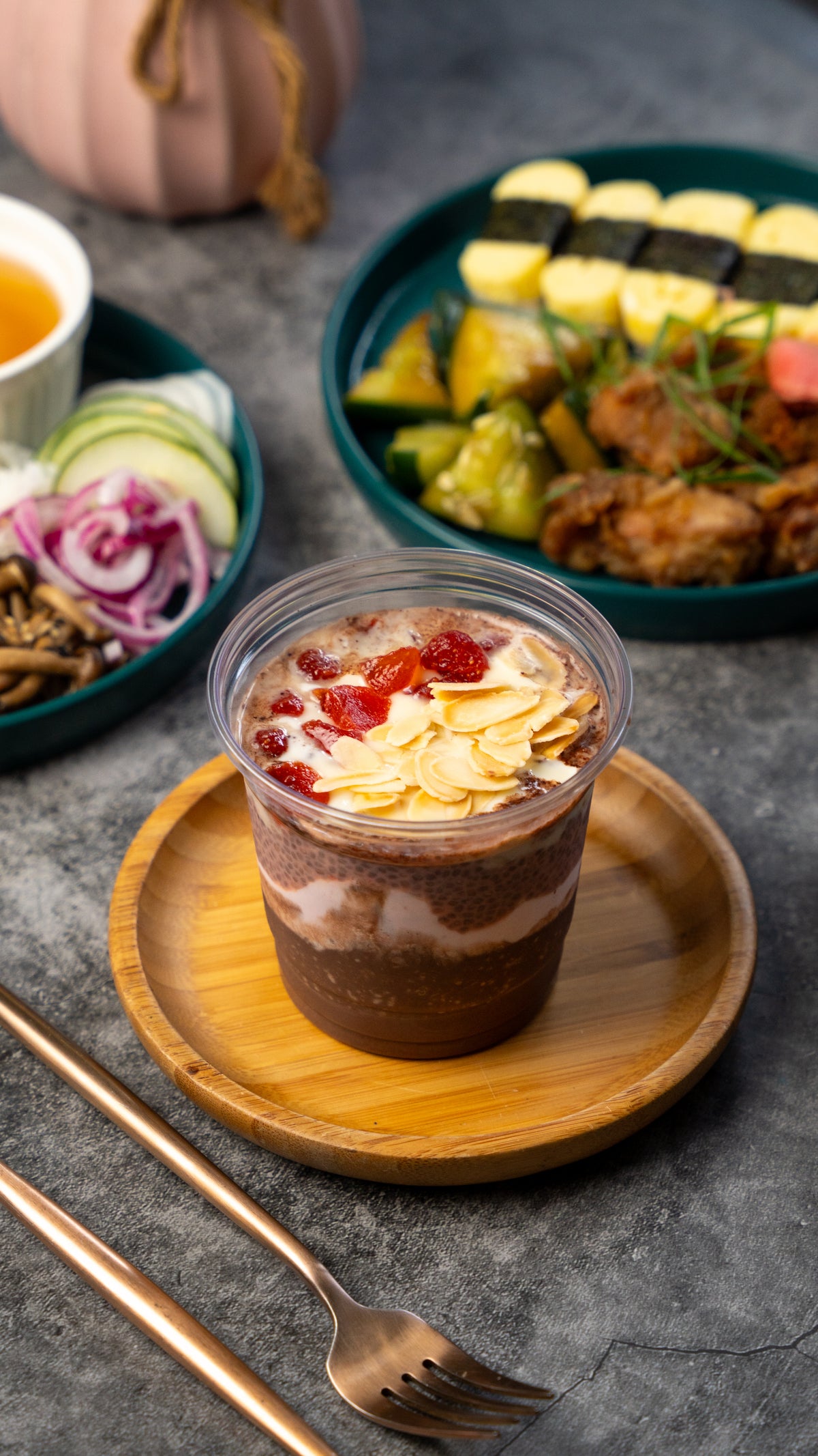 A clear plastic cup of layered dessert or iced chocolate pudding topped with sliced almonds and red fruit pieces, placed on a round wooden plate. The cup is set on a gray stone-like surface with a fork and spoon in the foreground, and bowls of sushi and other dishes blurred in the background.