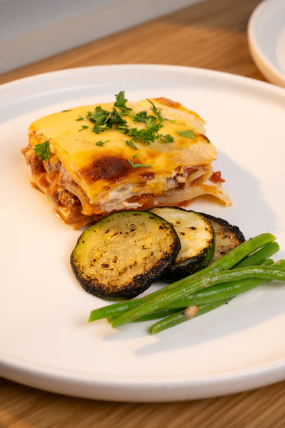 A plated slice of cheesy lasagna with green herb garnish, served with two grilled zucchini slices and green beans on a white plate over a wooden table.