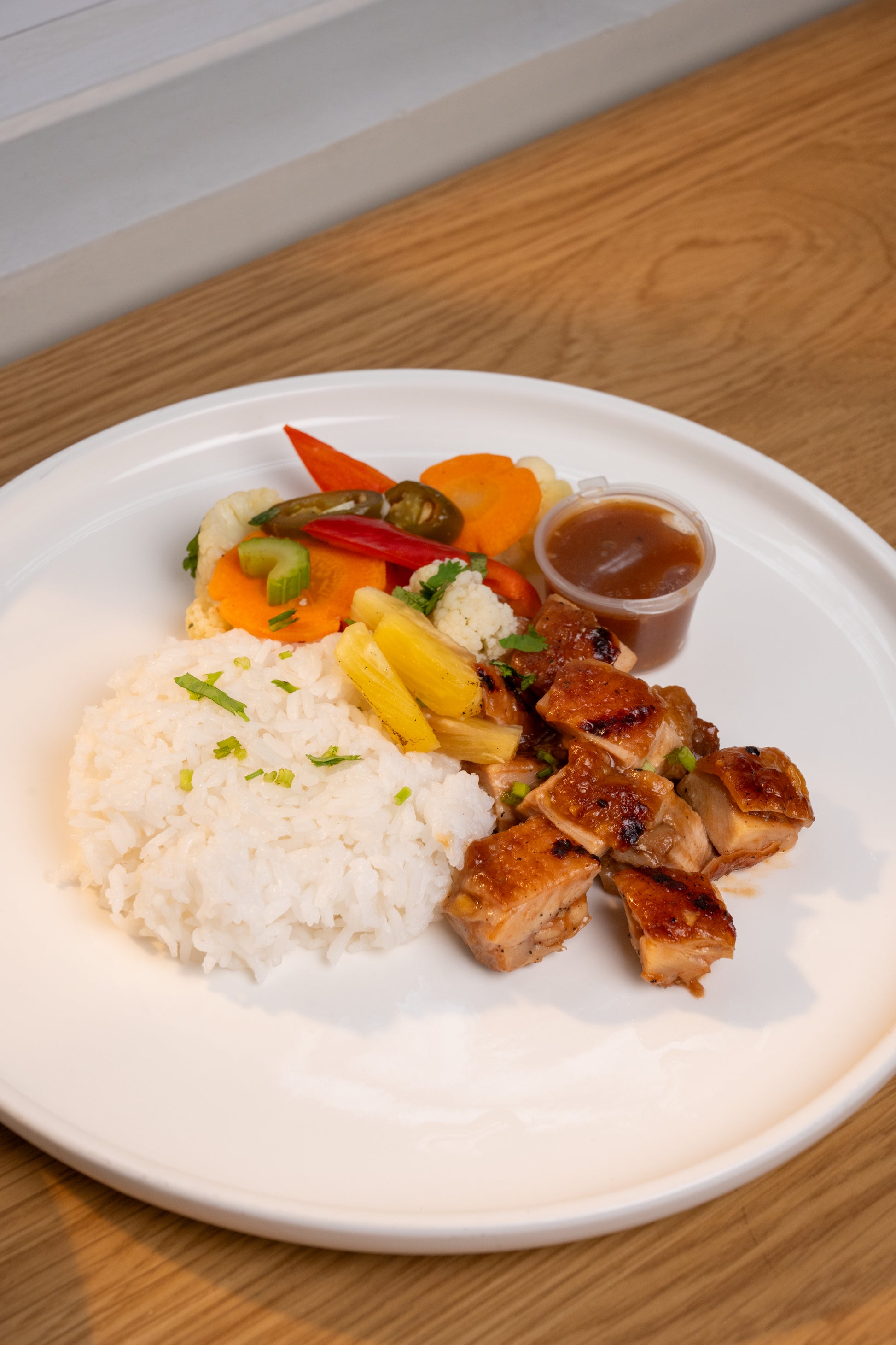 A plated meal on a white plate featuring glazed chicken pieces, a mound of white rice, mixed vegetables, and a small cup of brown sauce, set on a wooden table.