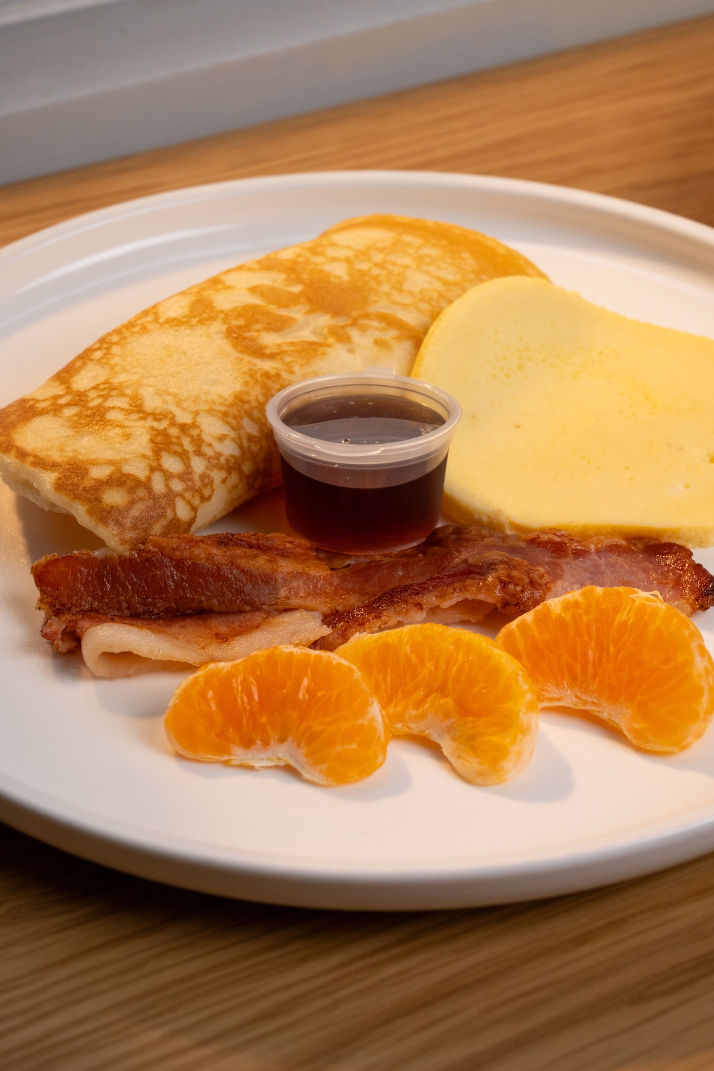 A breakfast plate featuring a folded crepe, a slice of yellow cheese, strips of bacon, a small cup of dark syrup, and three orange segments on a white plate, set on a wooden table.