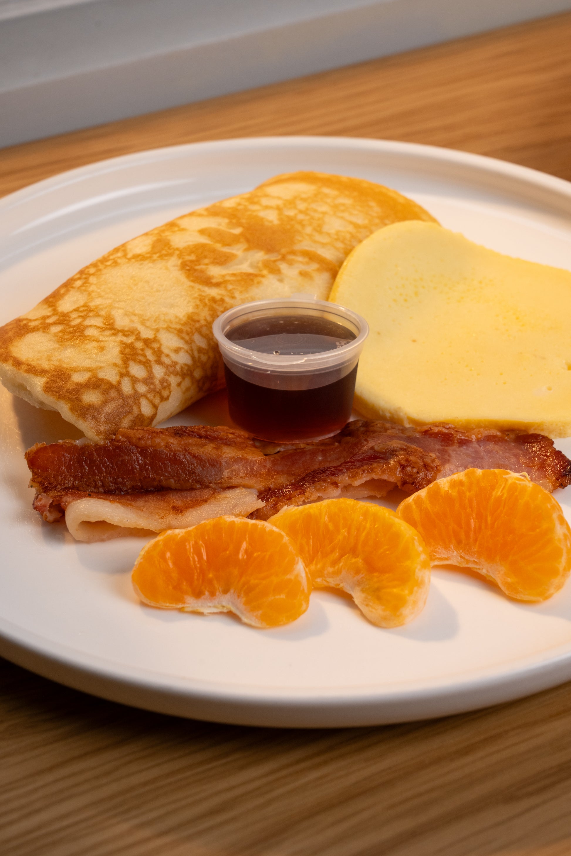 A breakfast plate featuring a folded crepe, a slice of yellow cheese, strips of bacon, a small cup of dark syrup, and three orange segments on a white plate, set on a wooden table.