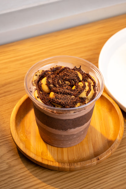 A clear plastic cup filled with layered chocolate dessert and topped with a dark chocolate swirl, nuts, and banana pieces, sitting on a round wooden tray on a wooden table.