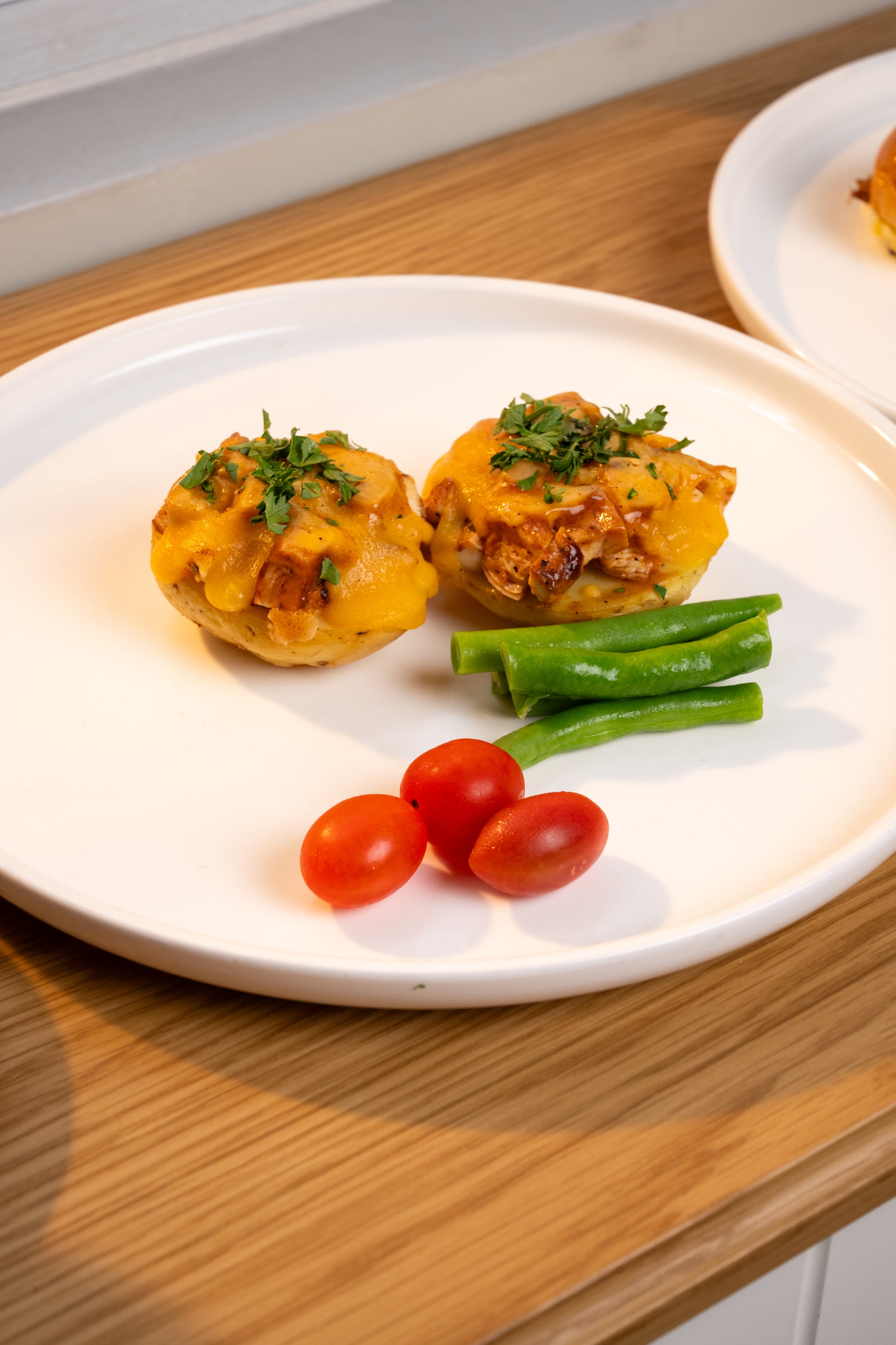 Two baked potato cups topped with melted cheese and chopped green herbs, served on a white plate with green beans and cherry tomatoes on a wooden table.