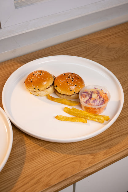 Two small sesame seed hamburger buns with golden-brown patties, served with two strips of french fries and a small clear cup of coleslaw on a large white plate, set on a wooden table.