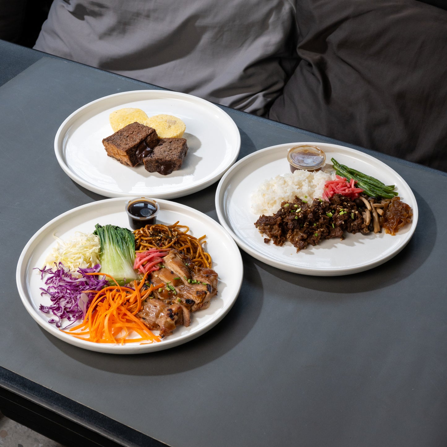Three white plates of prepared food arranged on a dark gray table, with a person's gray jacket visible in the background.
