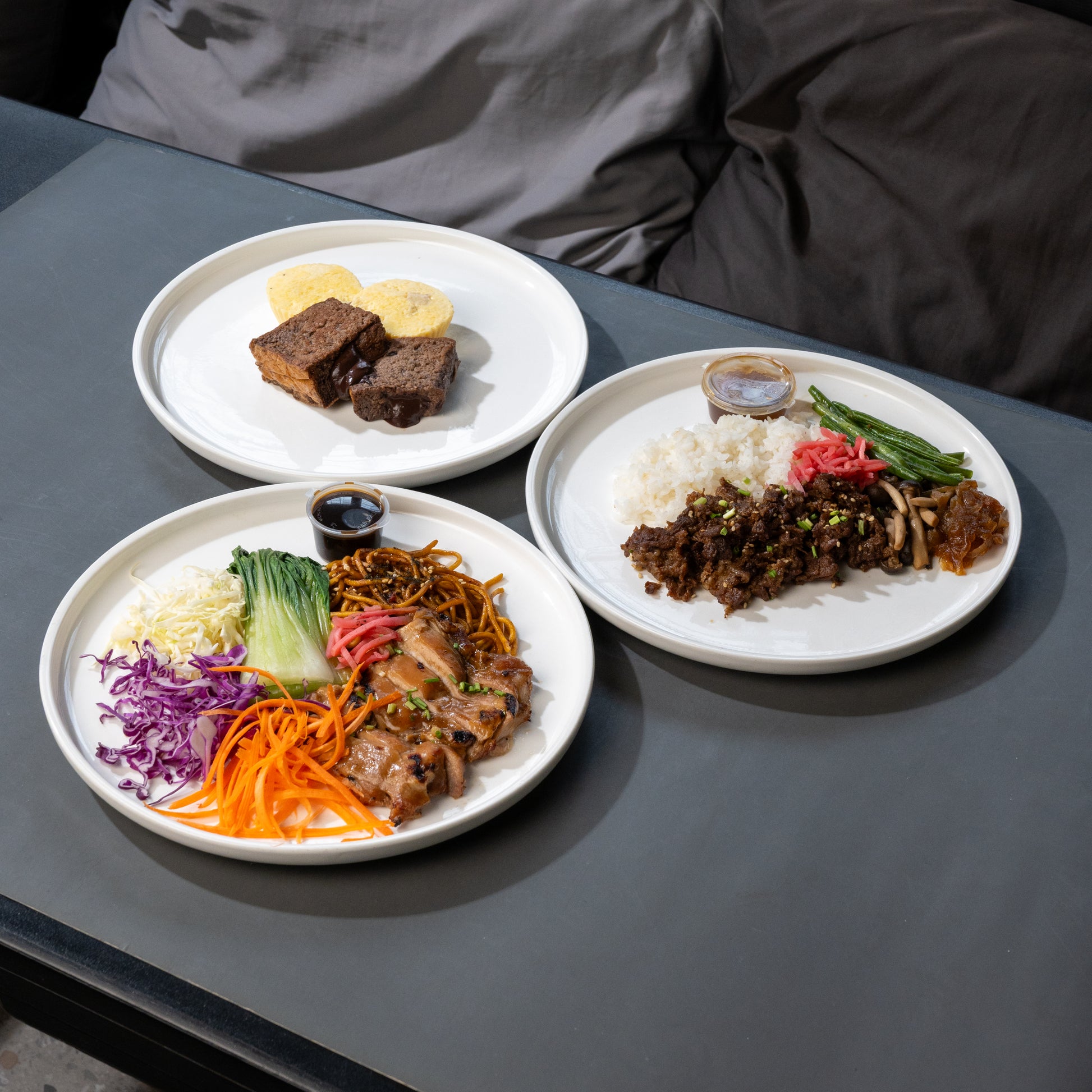 Three white plates of prepared food arranged on a dark gray table, with a person's gray jacket visible in the background.
