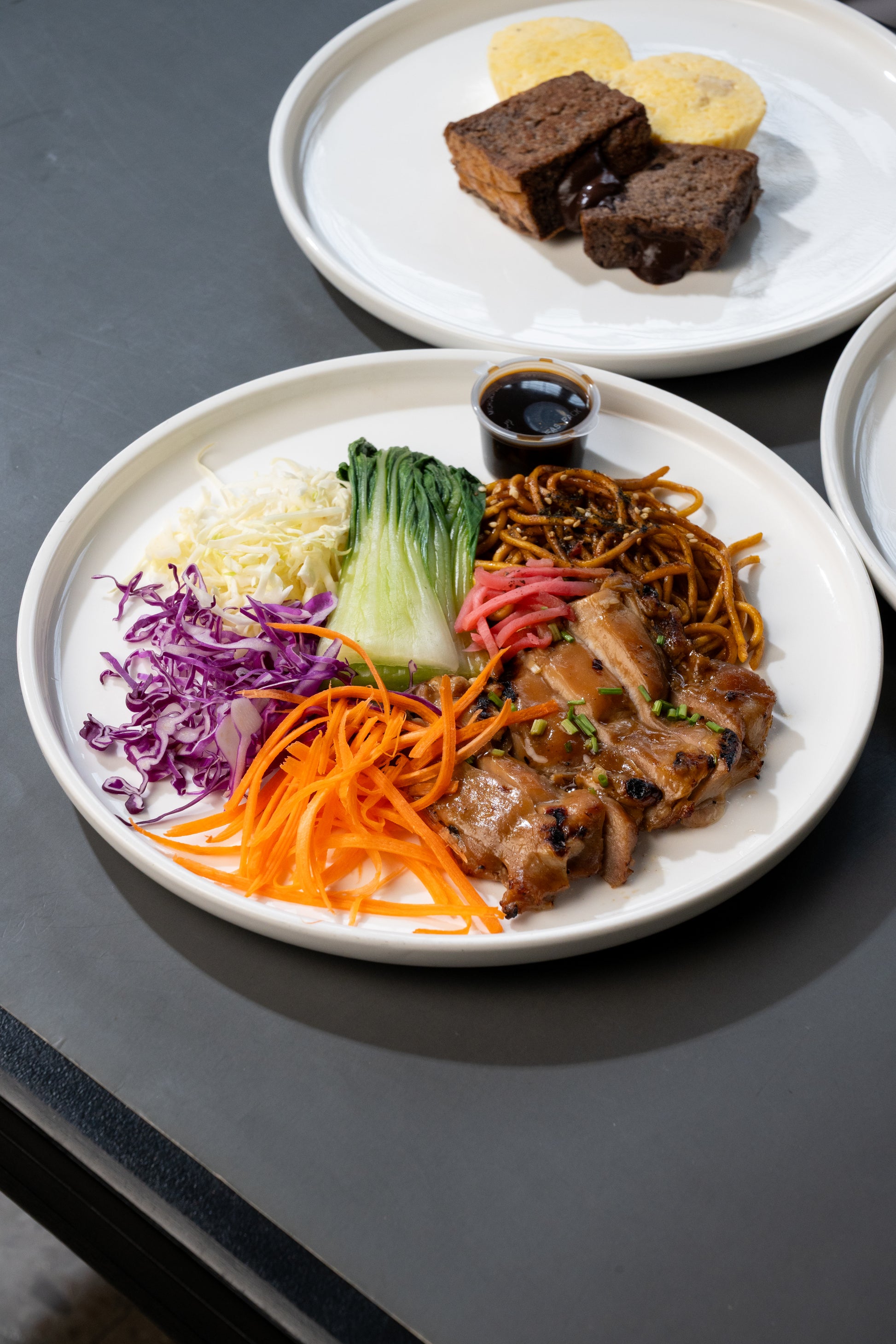 A plated dish of stir-fried chicken with noodles, shredded carrots, purple cabbage, white cabbage, and bok choy, served on a white plate on a dark gray table. Another plate in the background contains brown food items.