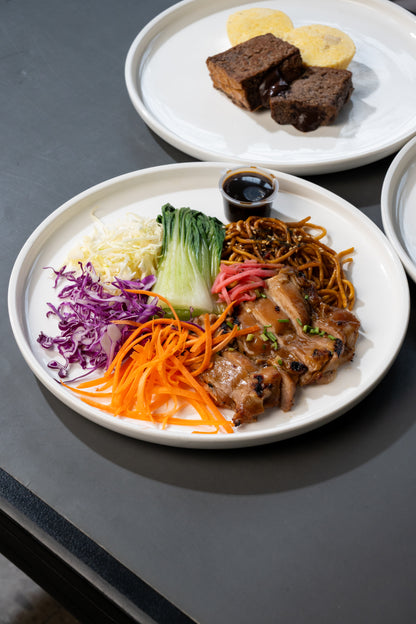 A plated dish of stir-fried chicken with noodles, shredded carrots, purple cabbage, white cabbage, and bok choy, served on a white plate on a dark gray table. Another plate in the background contains brown food items.