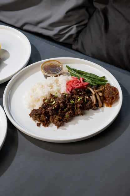 A plated meal on a dark tabletop, featuring white rice, a brown meat dish topped with green onions, green beans, pickled red ginger, and a small dipping sauce. A gray fabric surface is visible in the background.