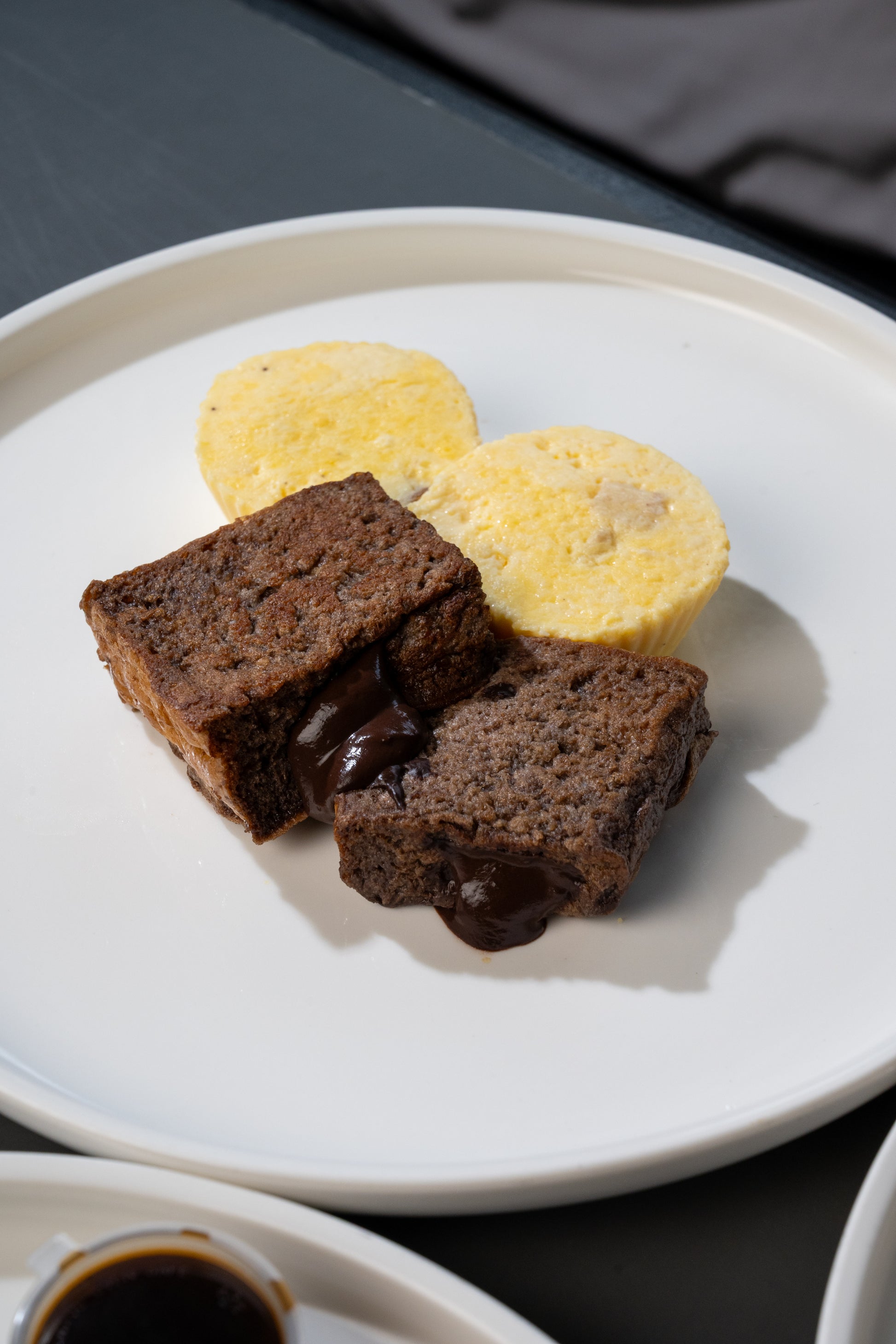 Two chocolate lava brownies with melted chocolate pools, served on a white plate with two yellow custard or cream puffs, set on a dark tabletop.