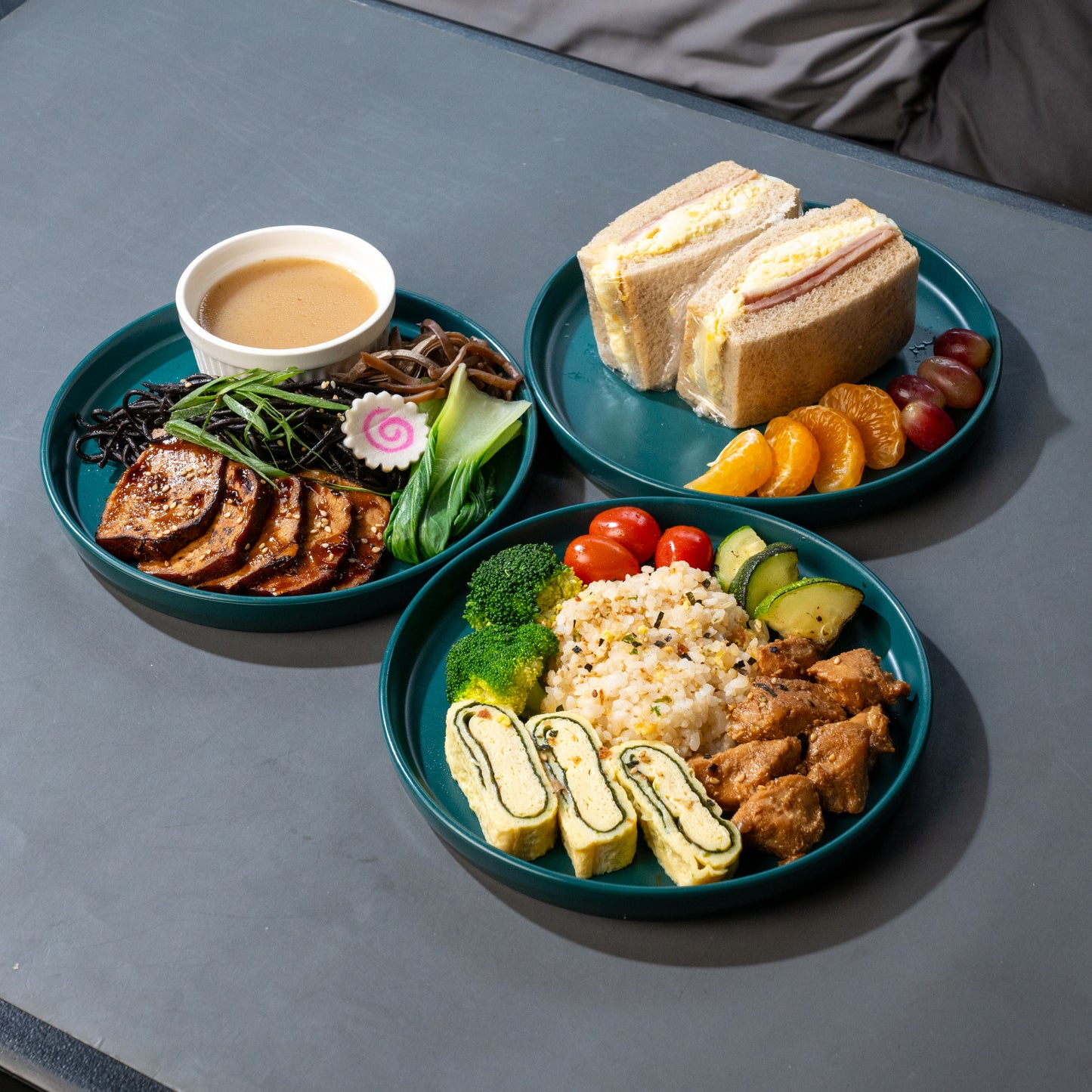 Three teal plates of food arranged on a dark gray table, including a sandwich with fruit, sliced grilled meat with sauce, and rice with vegetables and egg rolls.