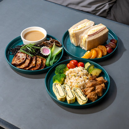 Three teal plates of food arranged on a dark gray table, including a sandwich with fruit, sliced grilled meat with sauce, and rice with vegetables and egg rolls.