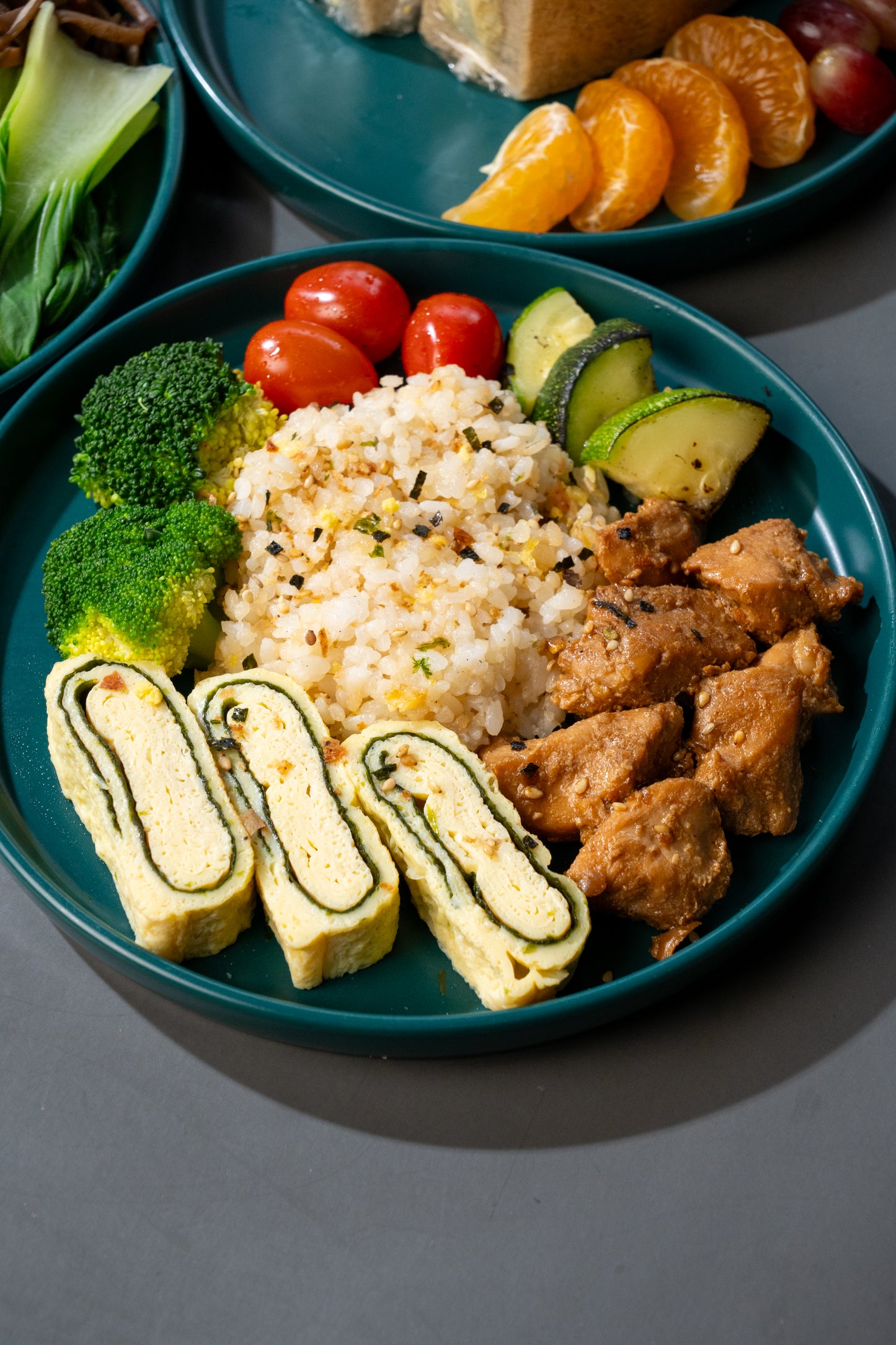 A close-up overhead shot of a teal plate filled with a rice-based snack or meal, including broccoli, cherry tomatoes, cucumber slices, rolled egg strips, and browned rectangular pieces, with partial fruit and another plate visible in the background.
