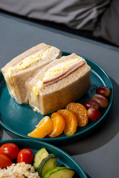 A close-up of a sandwich on a teal plate, served with sliced mandarin oranges, grapes, and a side bowl of salad in the foreground.
