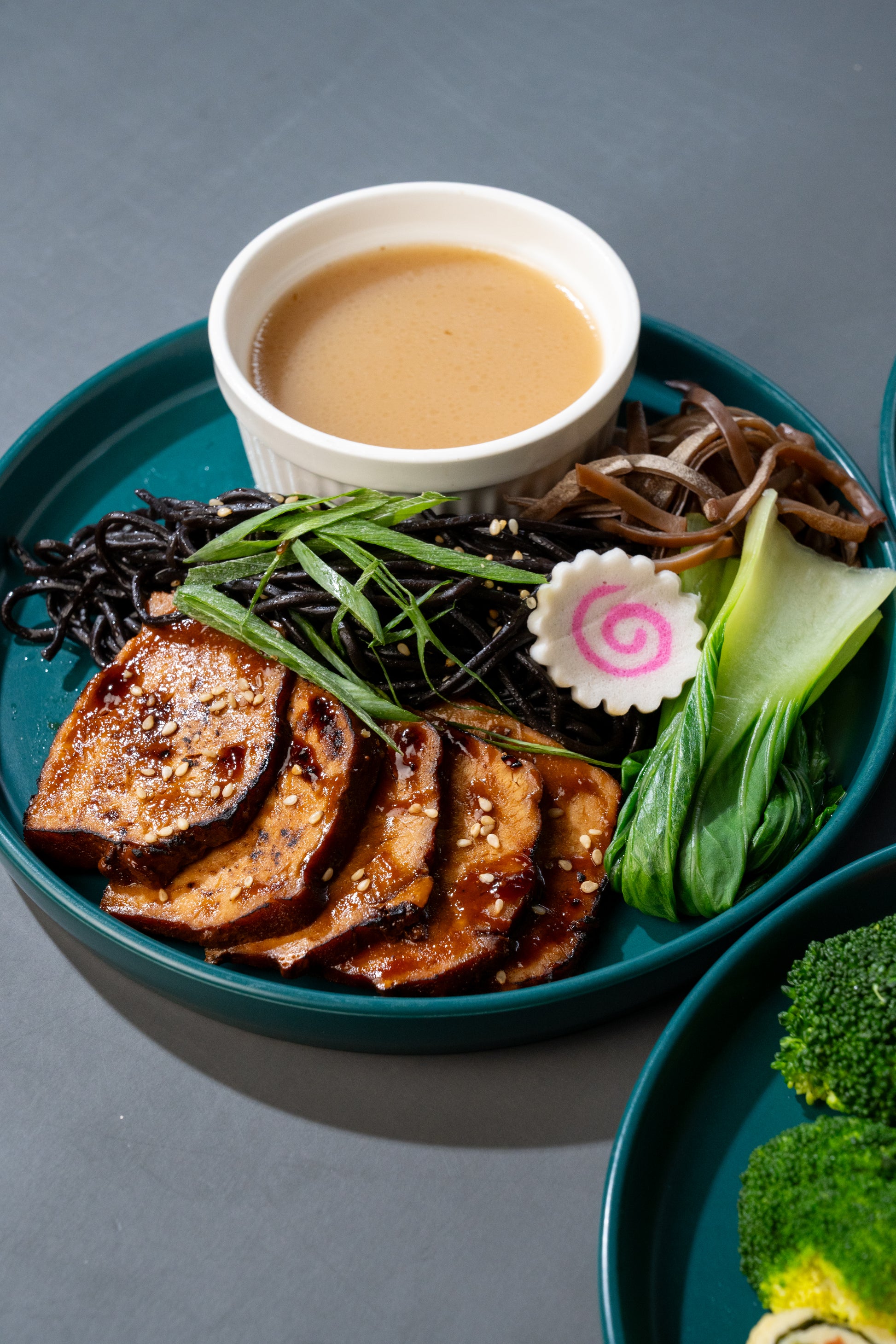 A plated prepared food dish on a dark teal plate, featuring glazed slices of tofu or tempeh, black seaweed, green onions, bok choy, mushrooms, and a small white bowl of light brown dipping sauce on a gray surface.